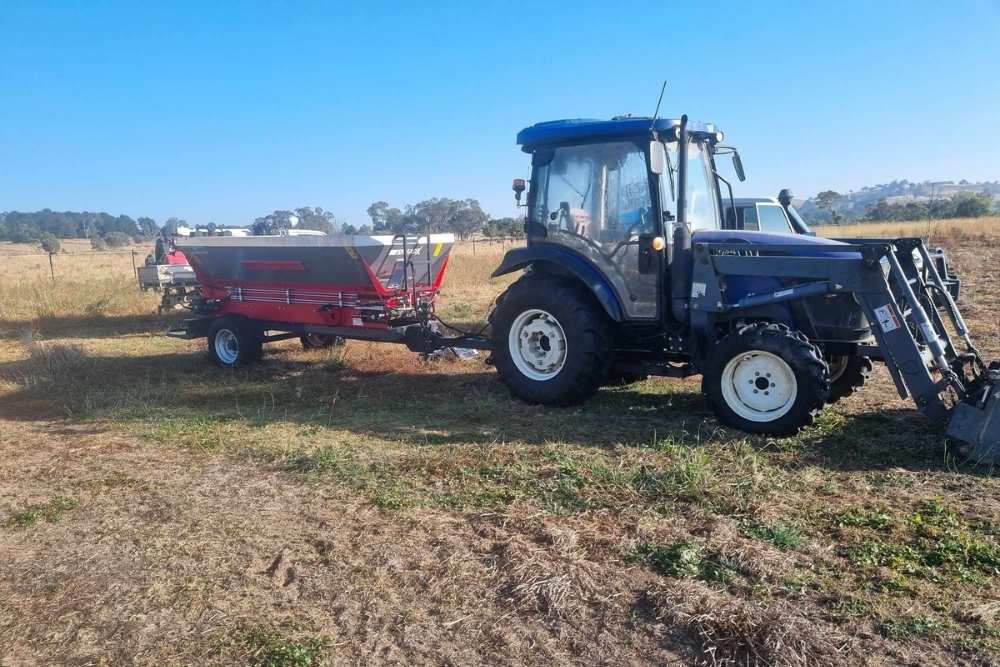 A tractor with a front end loader pulls a red and grey fertiliser spreader through a rural paddock-